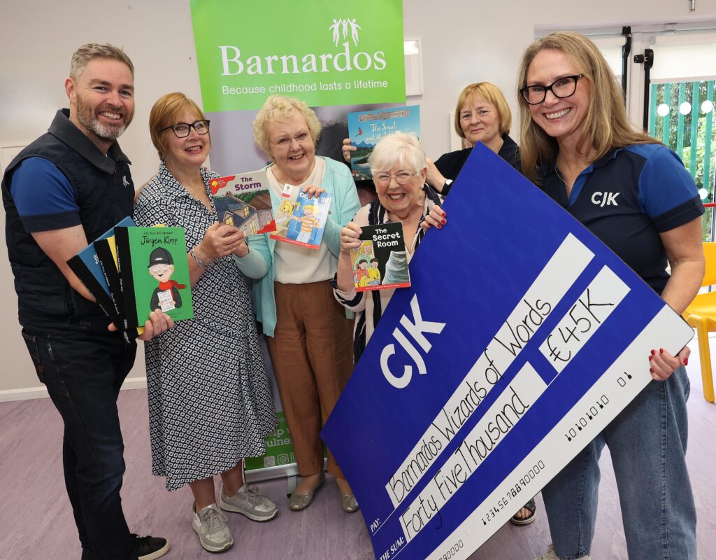 Six people stand in front of a Barnardos banner holding children’s books, while one person displays a large blue CJK charity cheque for €45,000. The group is gathered indoors in a bright room, with several books visible, including The Storm, The Secret Room, and Jürgen Klopp. The cheque text reads “Barnardos Wizards of Words” and “Forty Five Thousand €45K