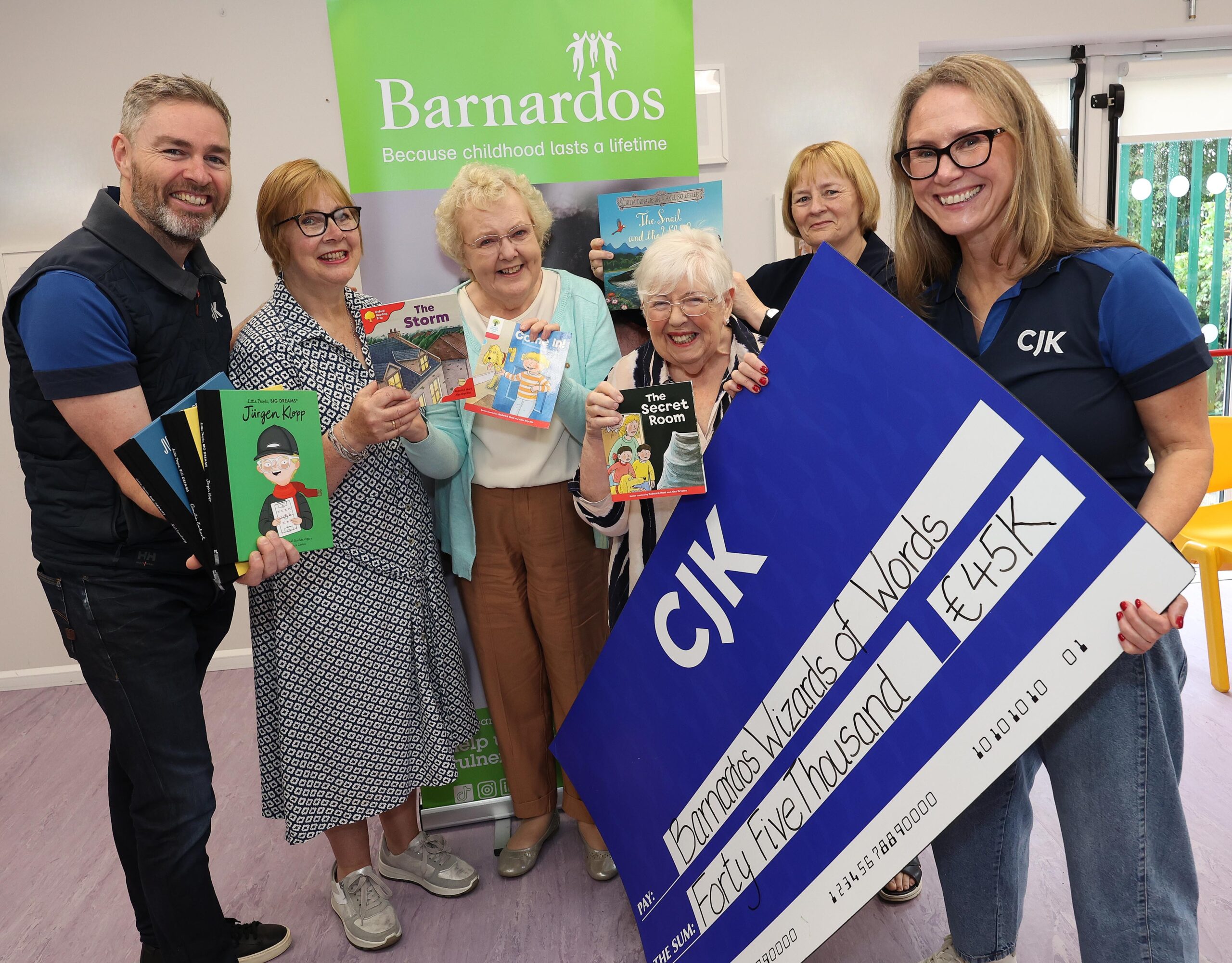 Six people stand in front of a Barnardos banner holding children’s books, while one person displays a large blue CJK charity cheque for €45,000. The group is gathered indoors in a bright room, with several books visible, including The Storm, The Secret Room, and Jürgen Klopp. The cheque text reads “Barnardos Wizards of Words” and “Forty Five Thousand €45K